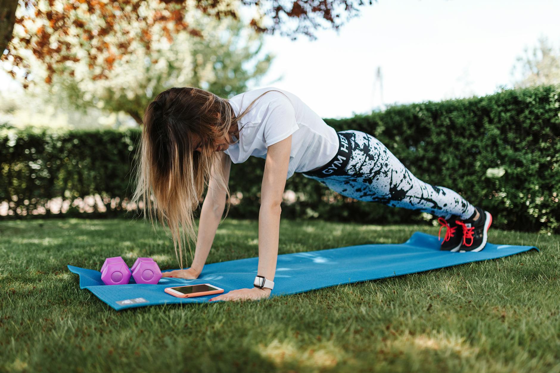 Woman doing push-ups on a yoga mat outside with dumbbells and a smartphone nearby. - outdoor hiit spring