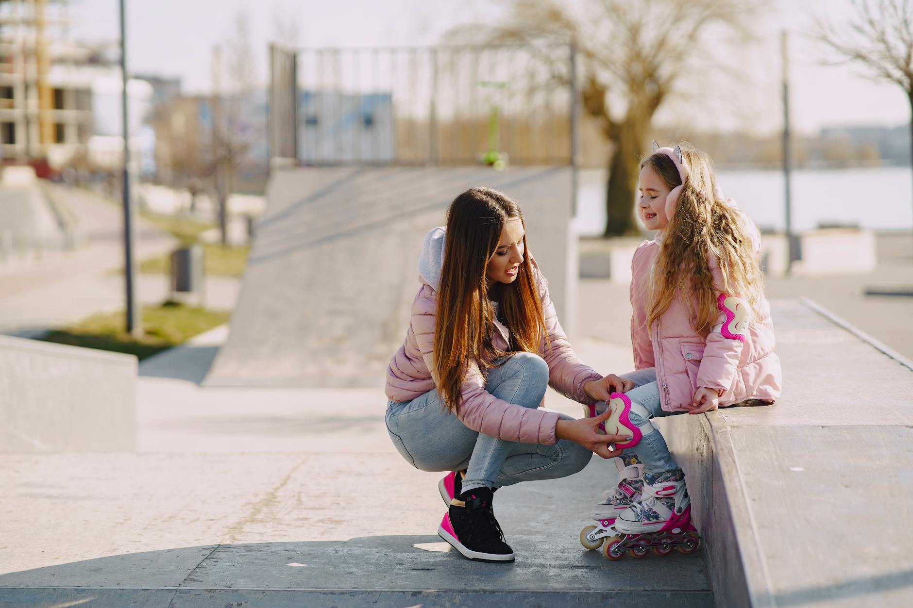 Cheerful young woman adjusting knee pads for little girl during active sport and game outside in city while spending time together - outdoor spring activities