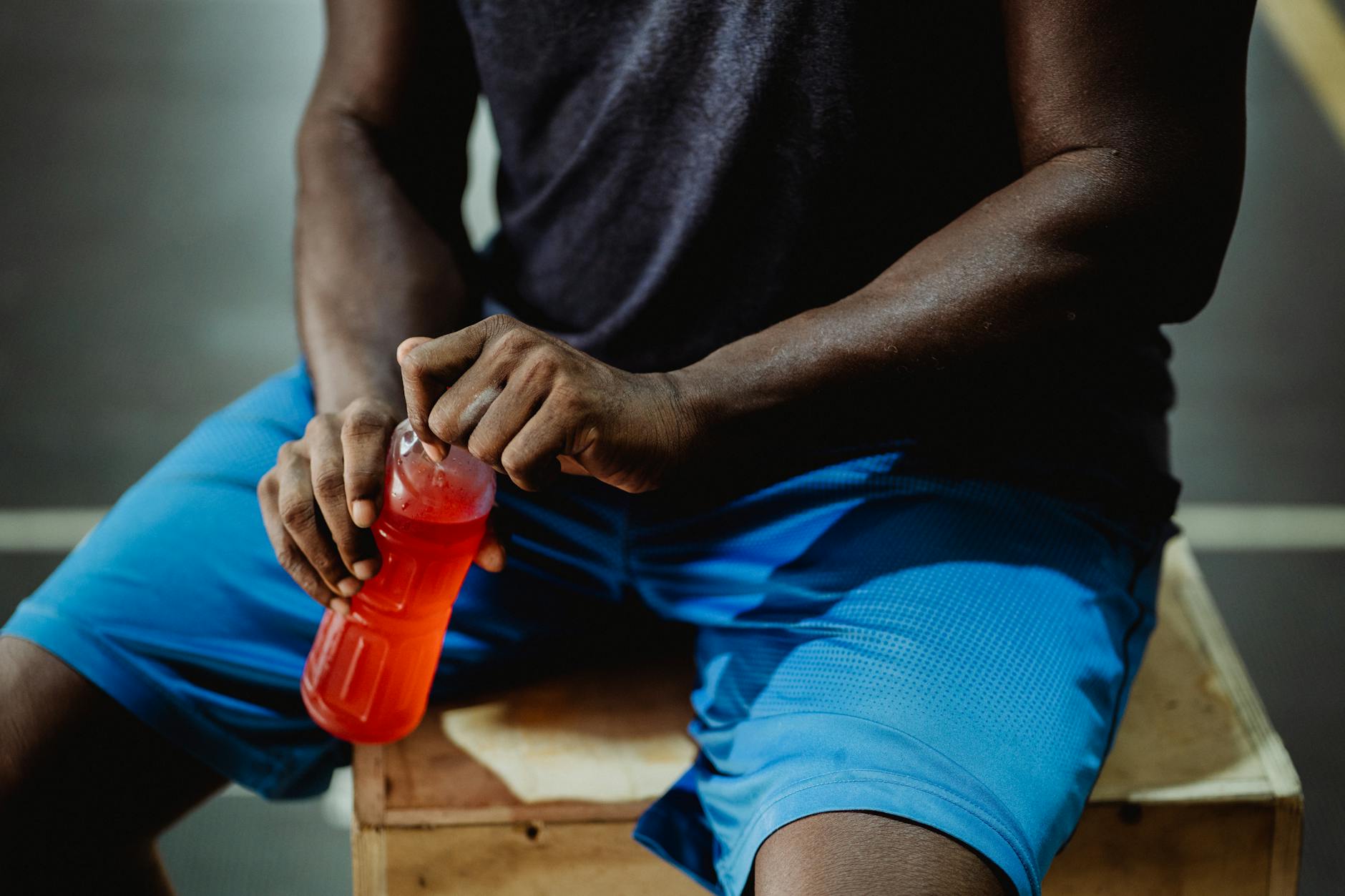 A man resting on a wooden box, enjoying a red sports drink during his workout session. - outdoor workout hydration
