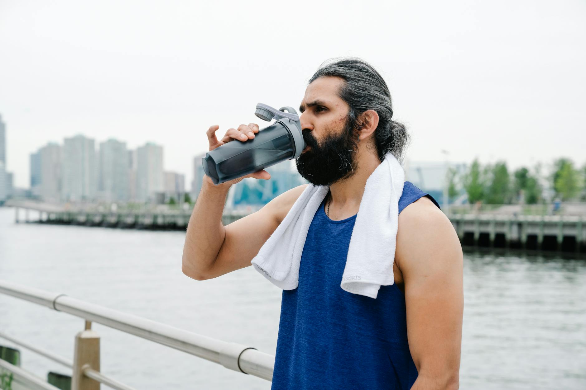 Man takes a refreshing drink by a riverside on a summer day with a cityscape background. - outdoor workout hydration
