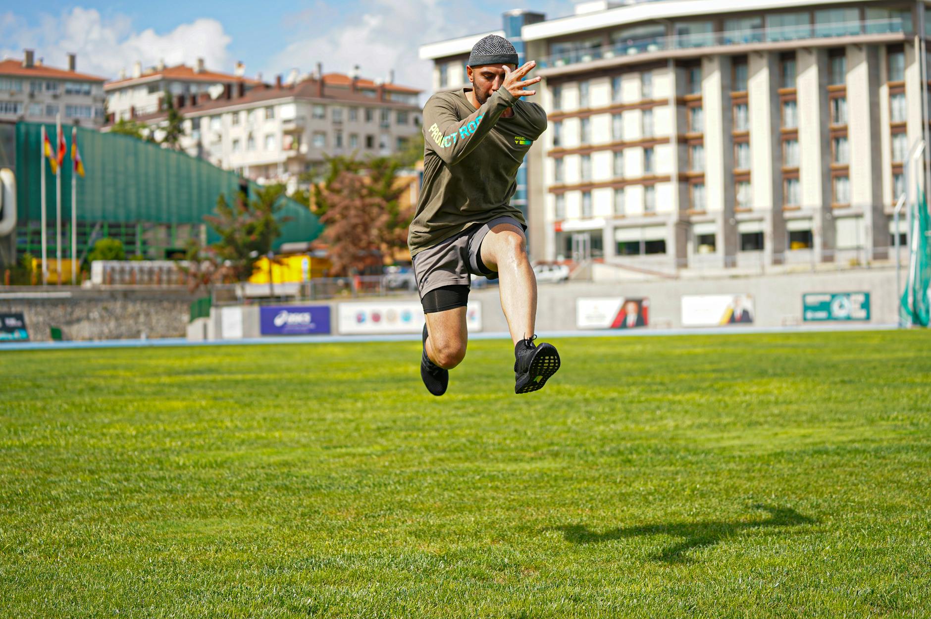 Energetic action photo of a man jumping on a grassy field in a cityscape background. - plyometric exercises