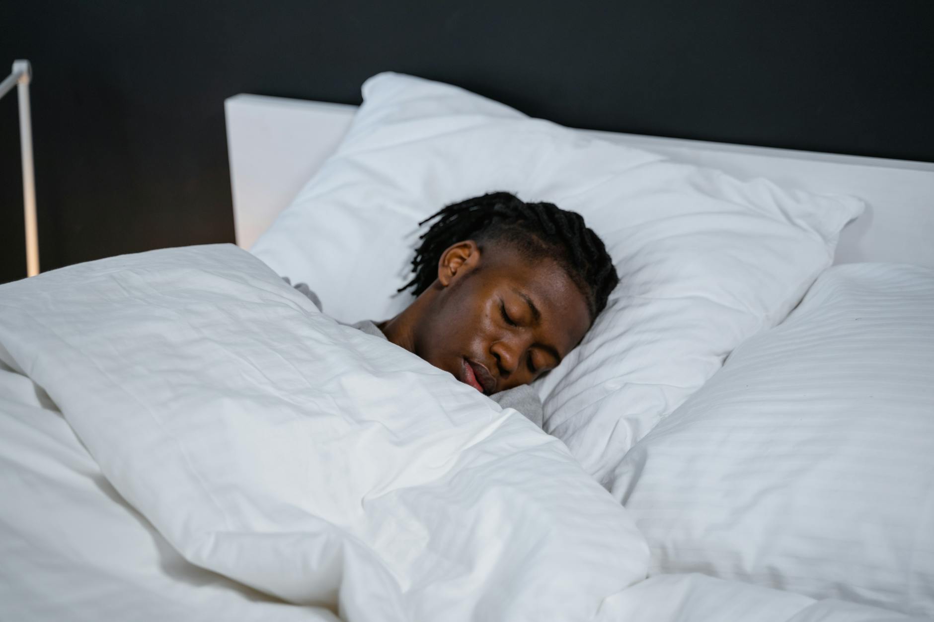 A young man resting peacefully in a bed with white sheets and a dark background. - sleep cycle reset