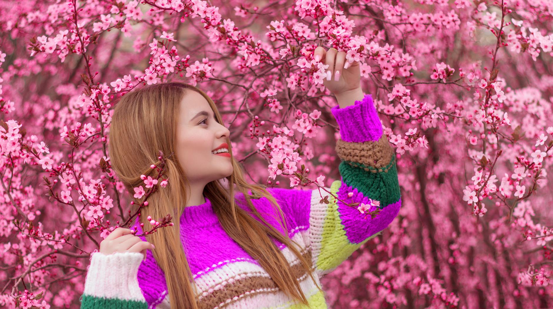A woman in a colorful sweater enjoying pink cherry blossoms outdoors, capturing spring's beauty. - spring allergy causes