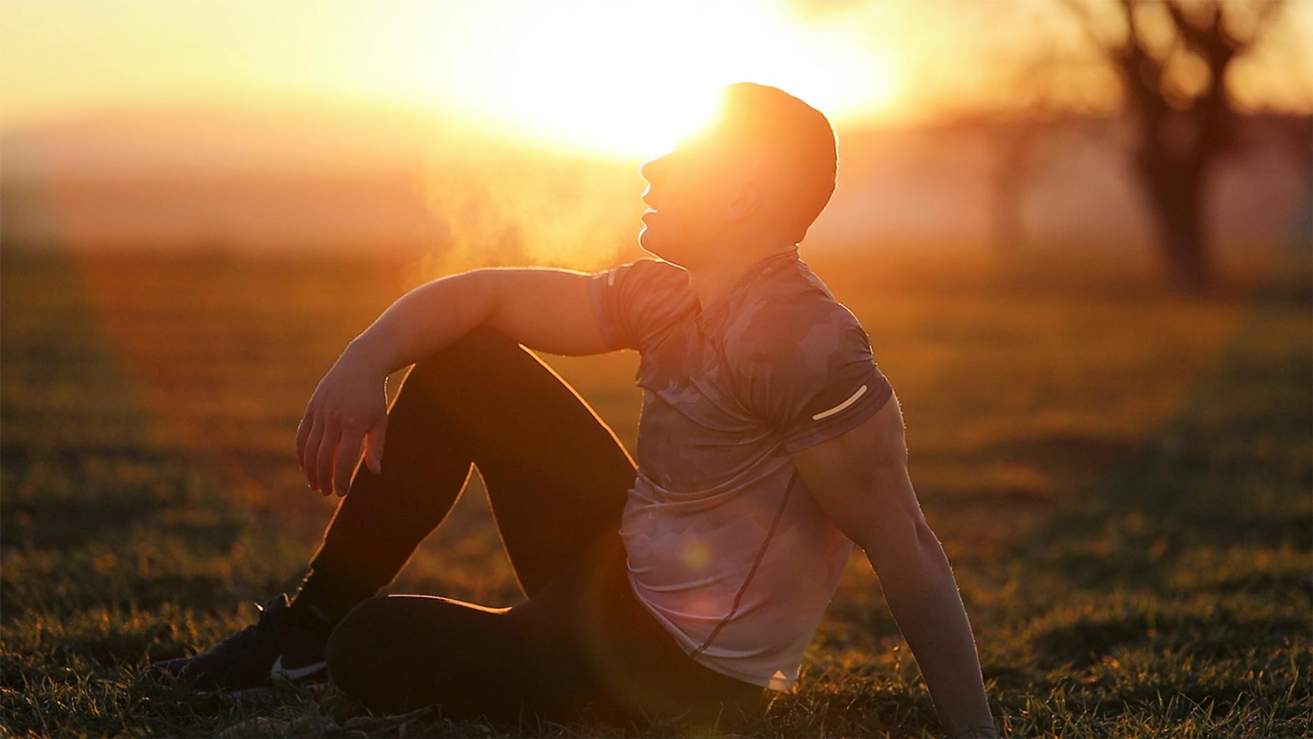 Muscular man sits in rural field at sunset, exhaling visible breath. - spring athlete recovery