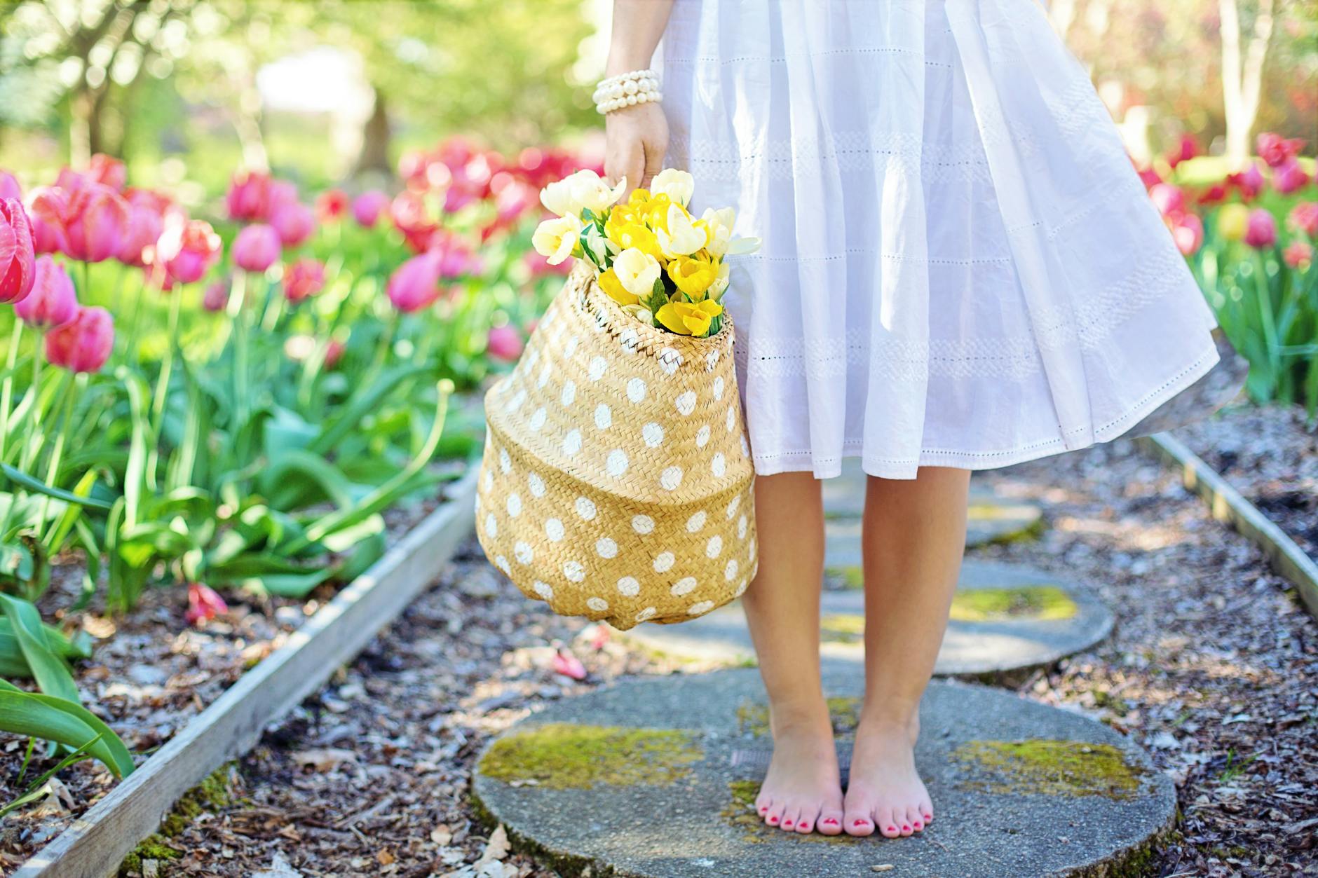 A woman in a white dress carries a basket of yellow flowers while walking barefoot in a tulip garden. - spring body reset
