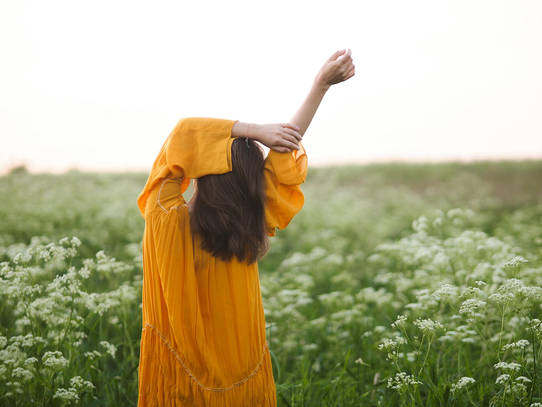 A woman in an orange dress with arms stretched, enjoying a field of wildflowers. - spring body reset