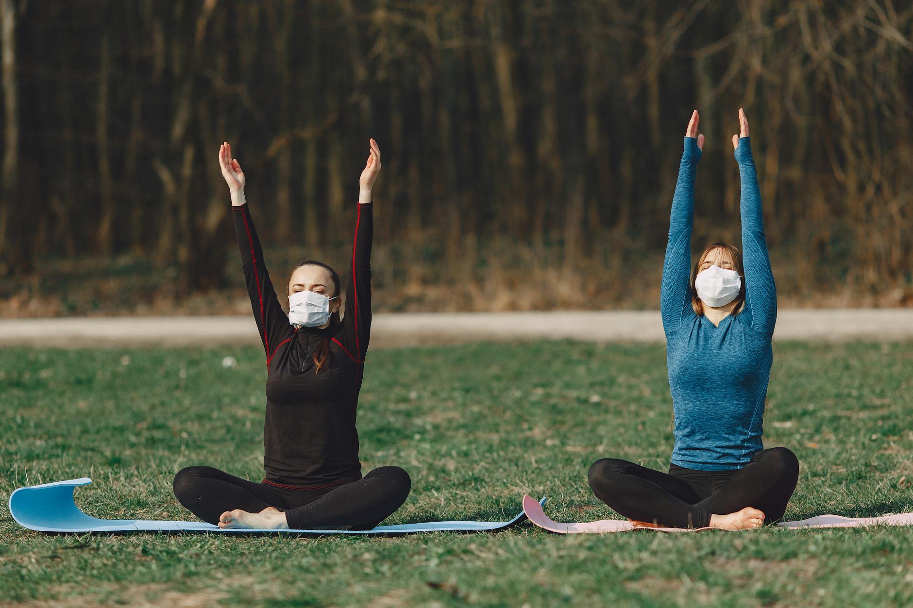 Slim female friends in sportswear and face masks sitting on mats with crossed legs and raised hands while practicing yoga with closed eyes in daylight during COVID 19 pandemic - spring exercise daylight