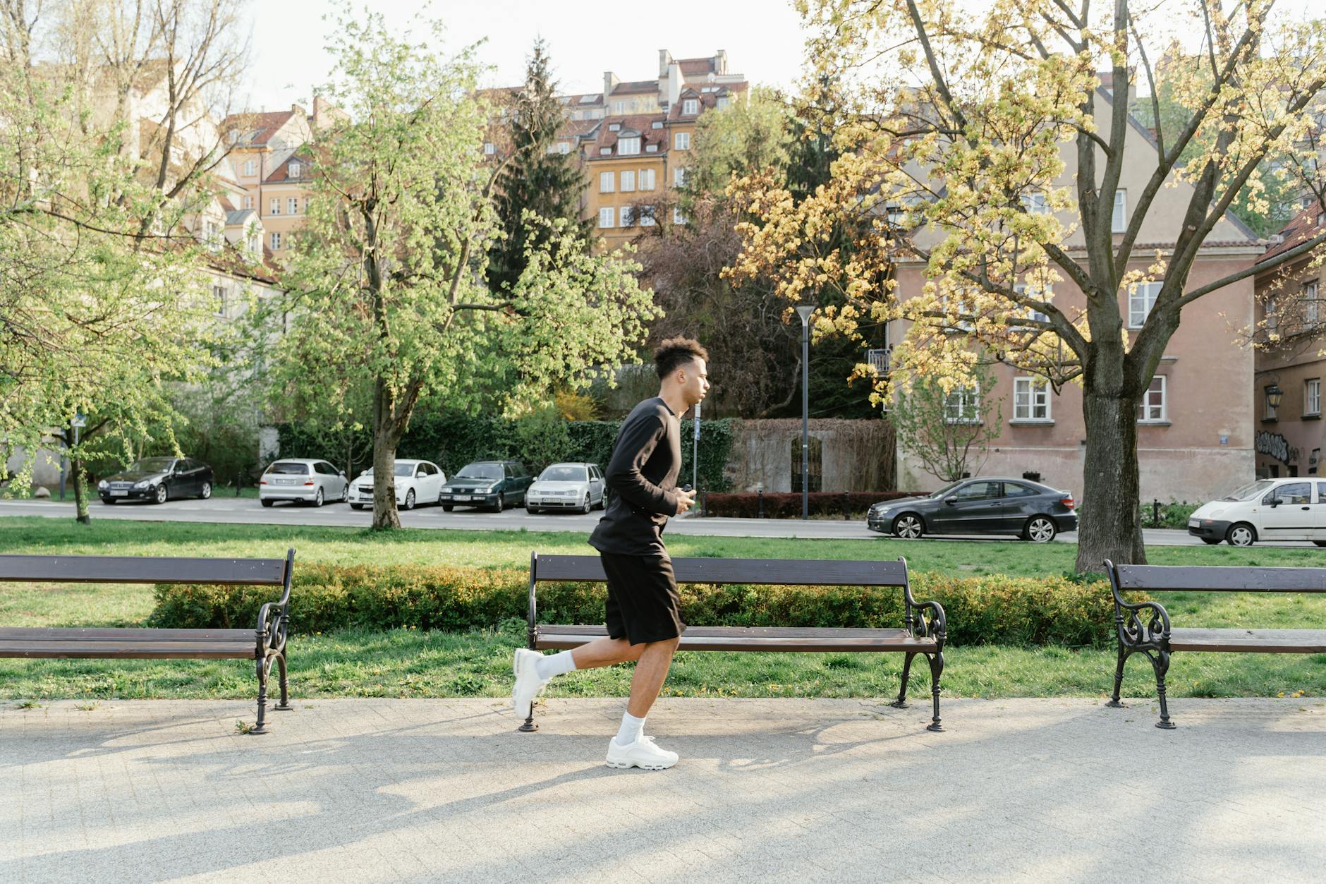 A man in athletic wear jogging through a sunlit park. Urban backdrop with trees and benches. - spring exercise mood