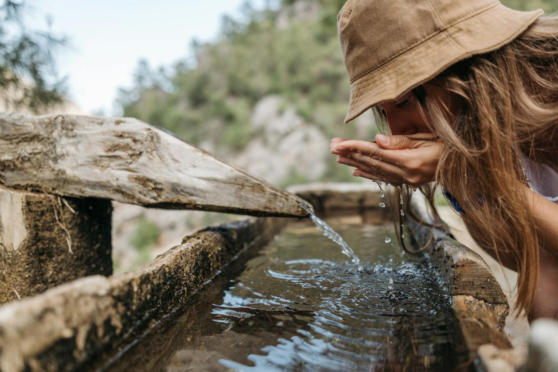 A young woman in a hat drinks from a rustic outdoor fountain in nature. - spring hike hydration