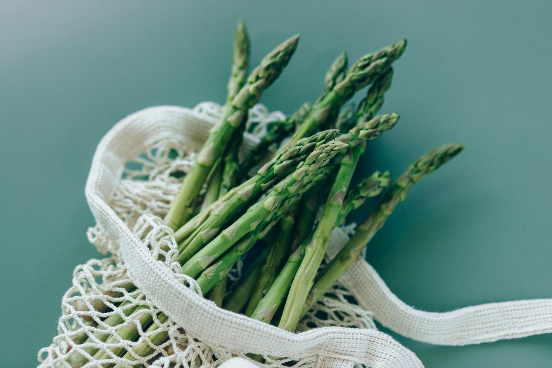 Fresh green asparagus in a reusable mesh bag on a green background, showcasing eco-friendly food storage. - spring mood foods