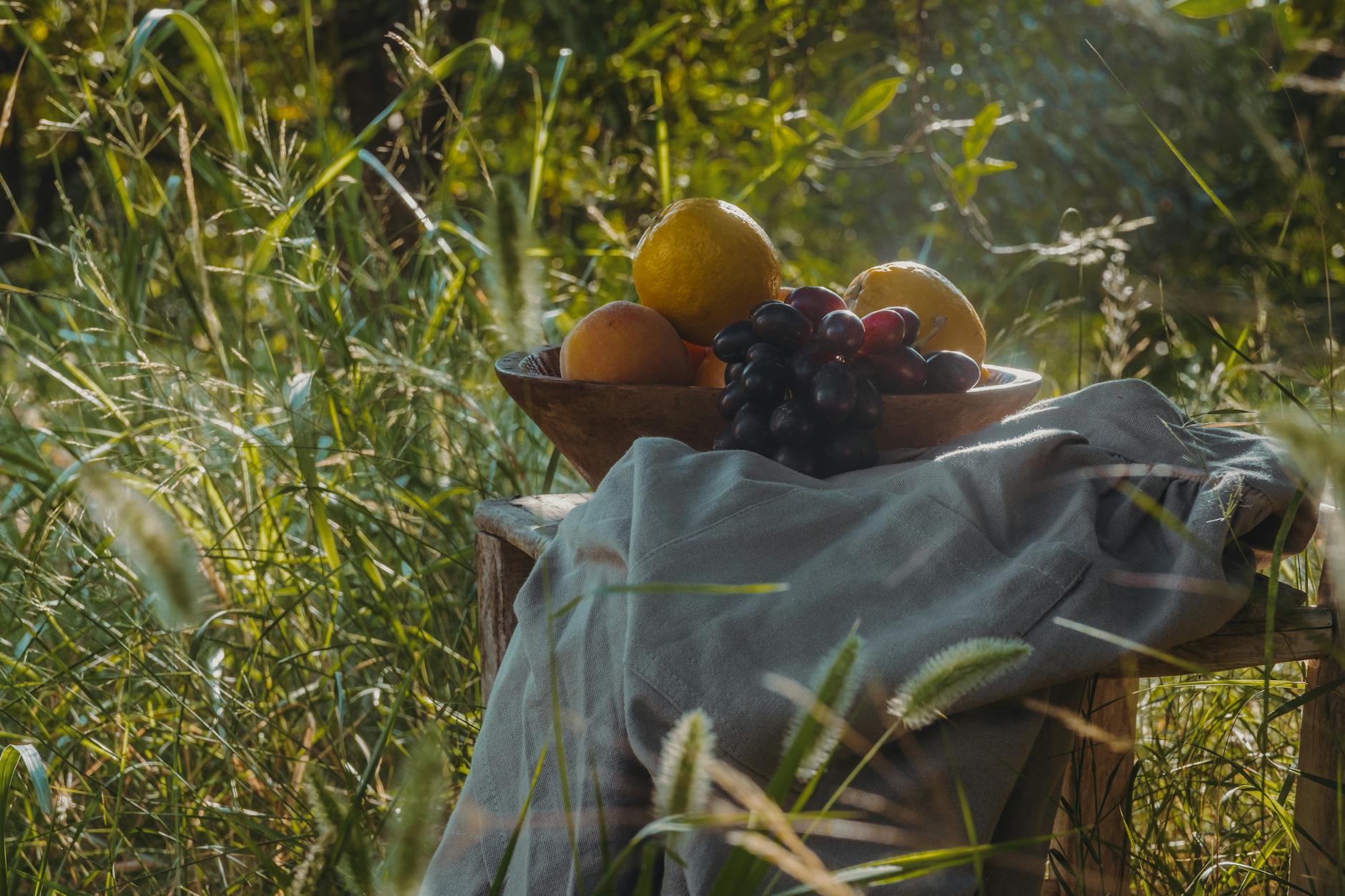 A rustic wooden bowl of fresh fruit including grapes and oranges in a sunlit field. - spring nutrition outdoor