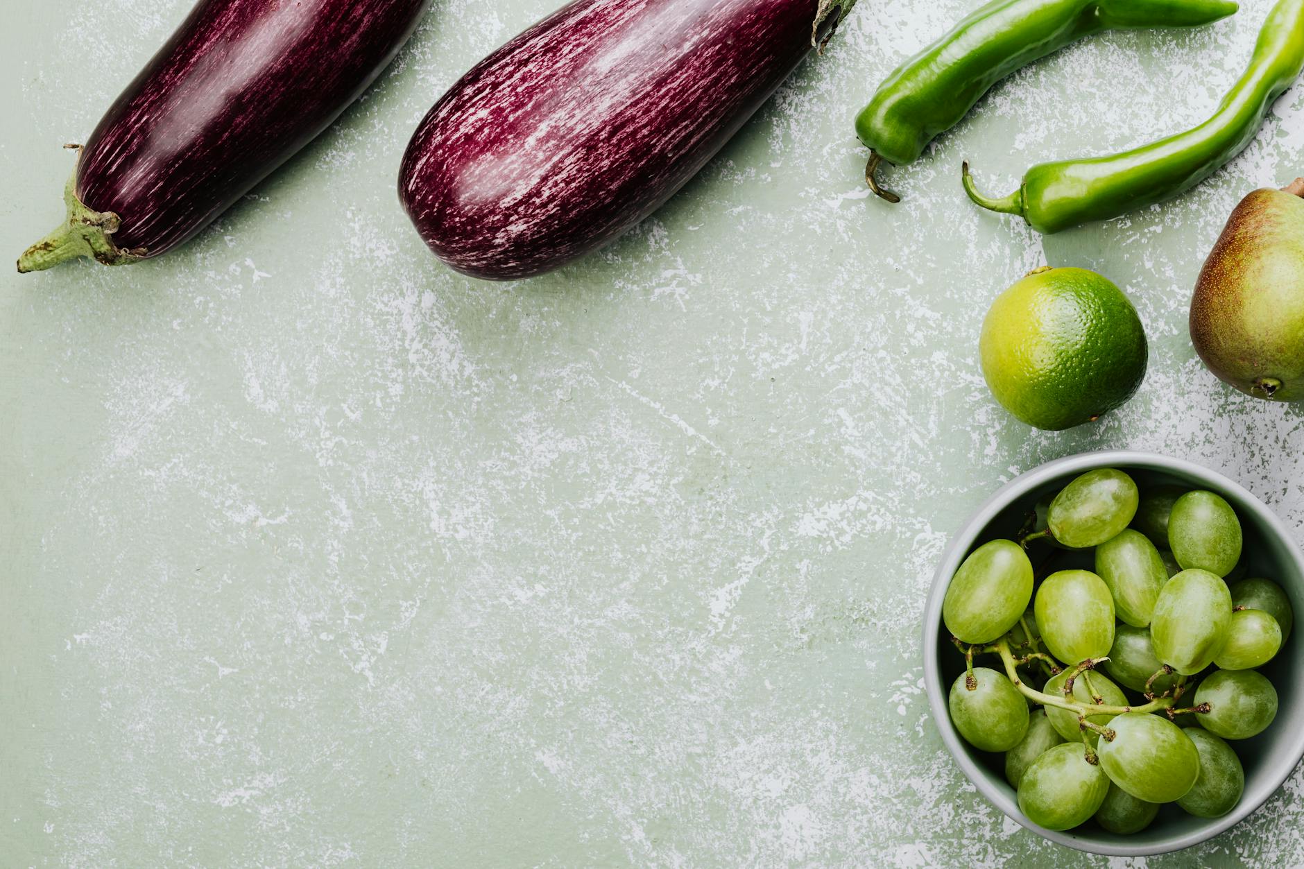 Top view of fresh vegetables and fruits featuring eggplants, green peppers, lime, pear, and grapes on a green surface. - spring produce recipes