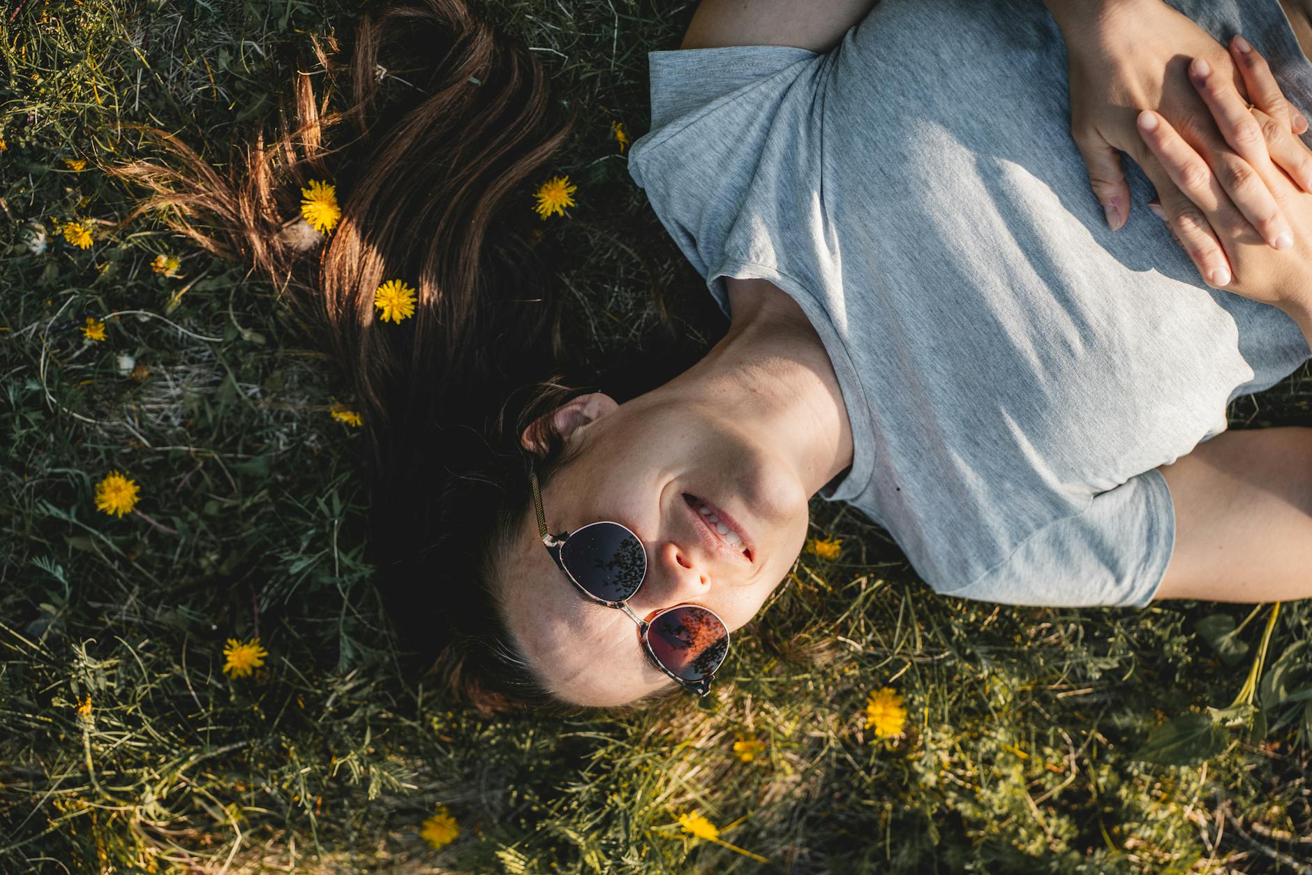 A woman enjoying a sunny day lying in a meadow full of dandelions in Québec, Canada wearing sunglasses. - spring self care