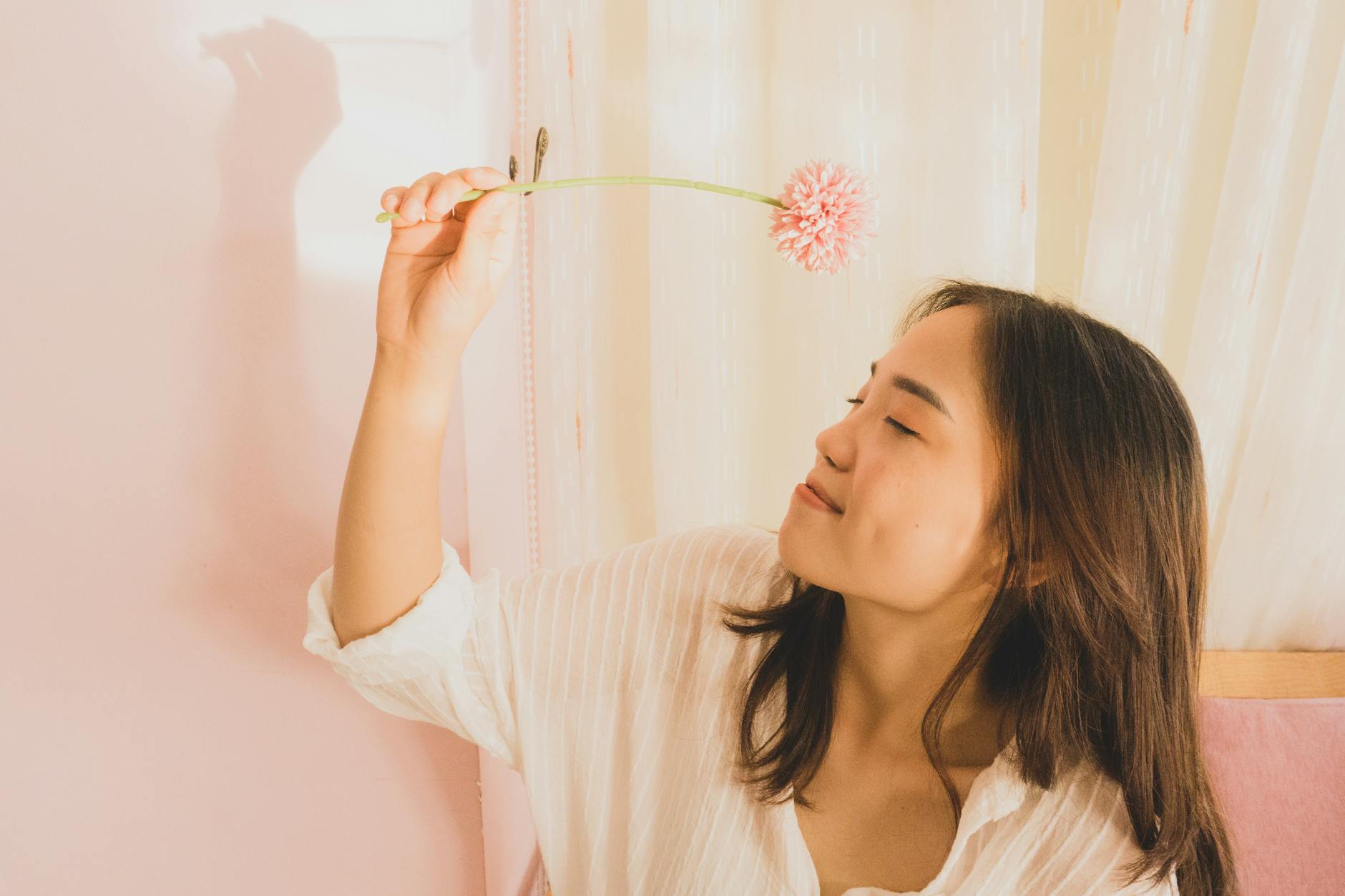 A serene portrait of a young woman holding a pink flower in a soft indoor setting. - spring self care