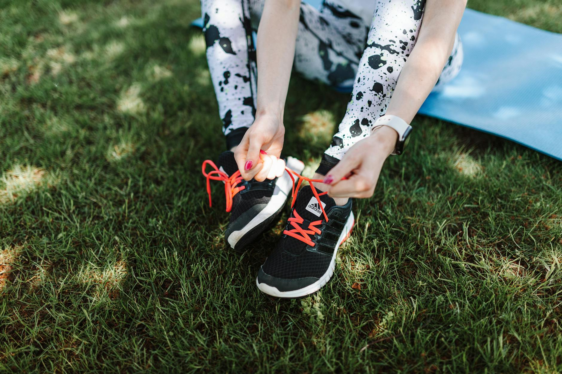 Woman tying Adidas sneakers on grass with yoga mat nearby, ready for exercise. - spring training gear