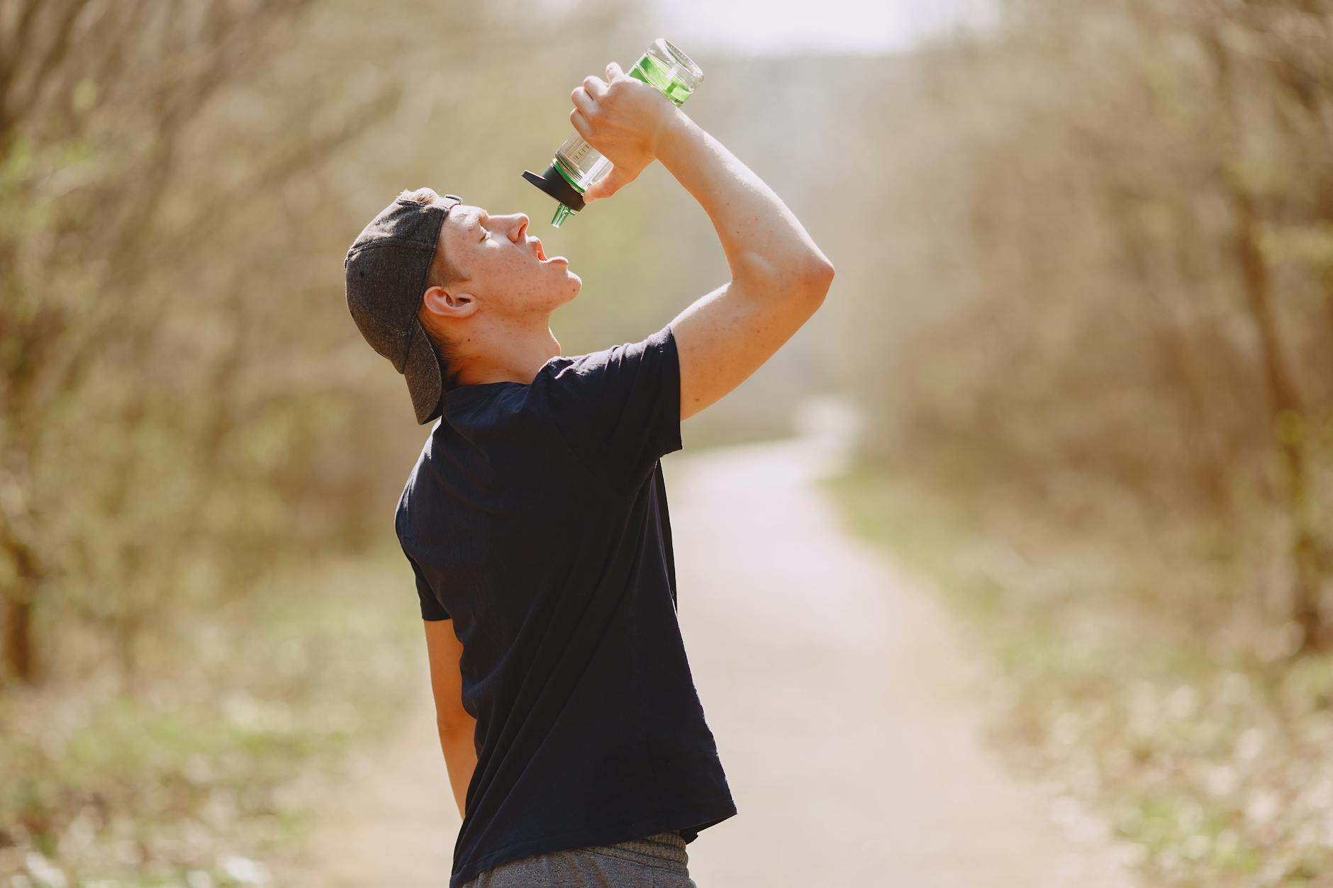 Side view of young male athlete wearing sports clothes and cap drinking water from plastic bottle on blurred background of forest during running and workout - spring training hydration