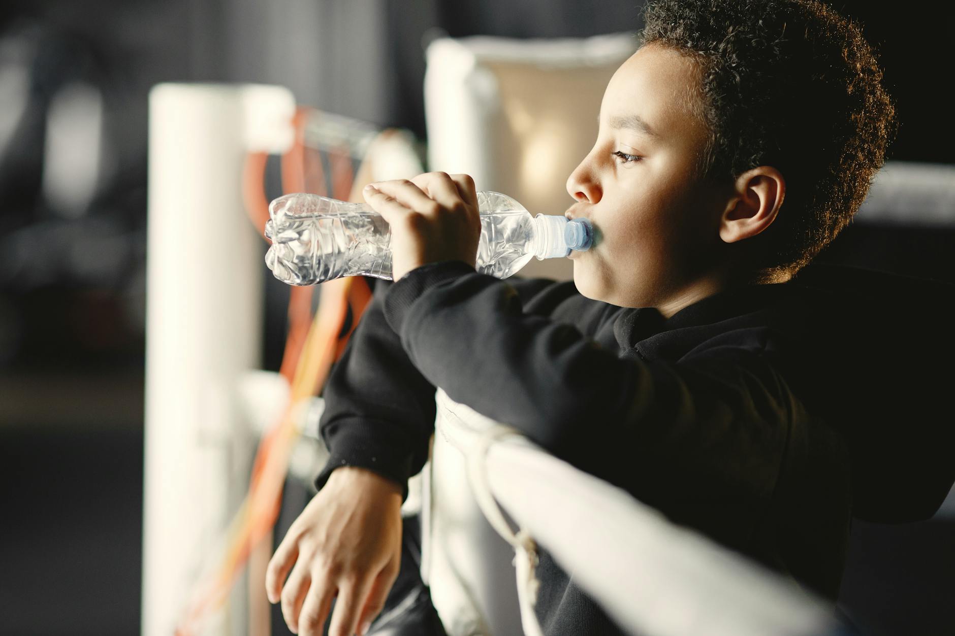 A young boy in a black hoodie drinks water from a plastic bottle in a boxing gym. - spring training hydration