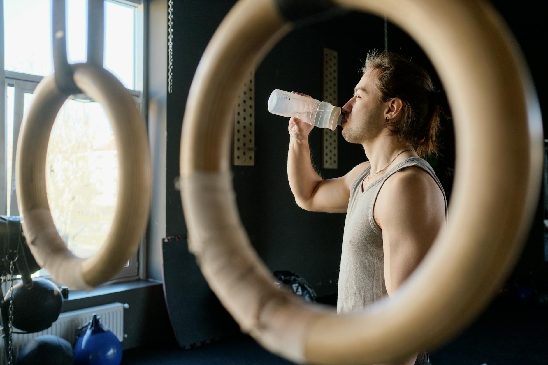 Athletic man drinking water in gym through gymnastic rings for hydration after workout. - spring training hydration