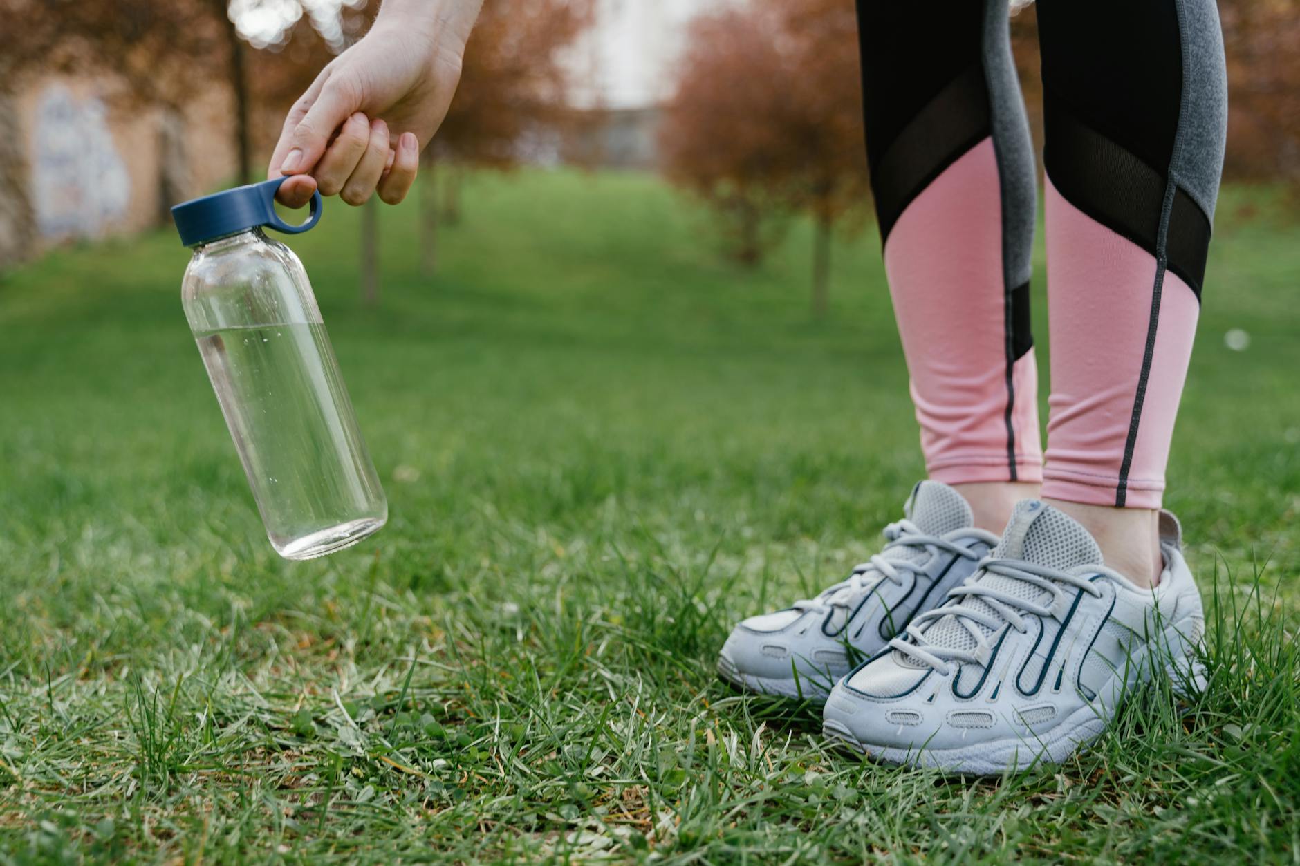 Close-up of legs in pink leggings and sneakers with a glass water bottle in hand on grass. - spring wellness habits