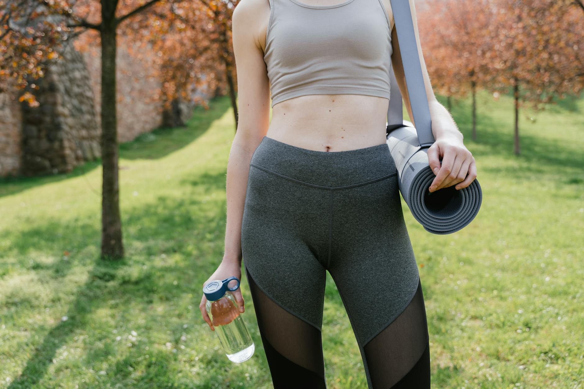 Woman in activewear holding yoga mat and water bottle in sunny park. - spring wellness routines