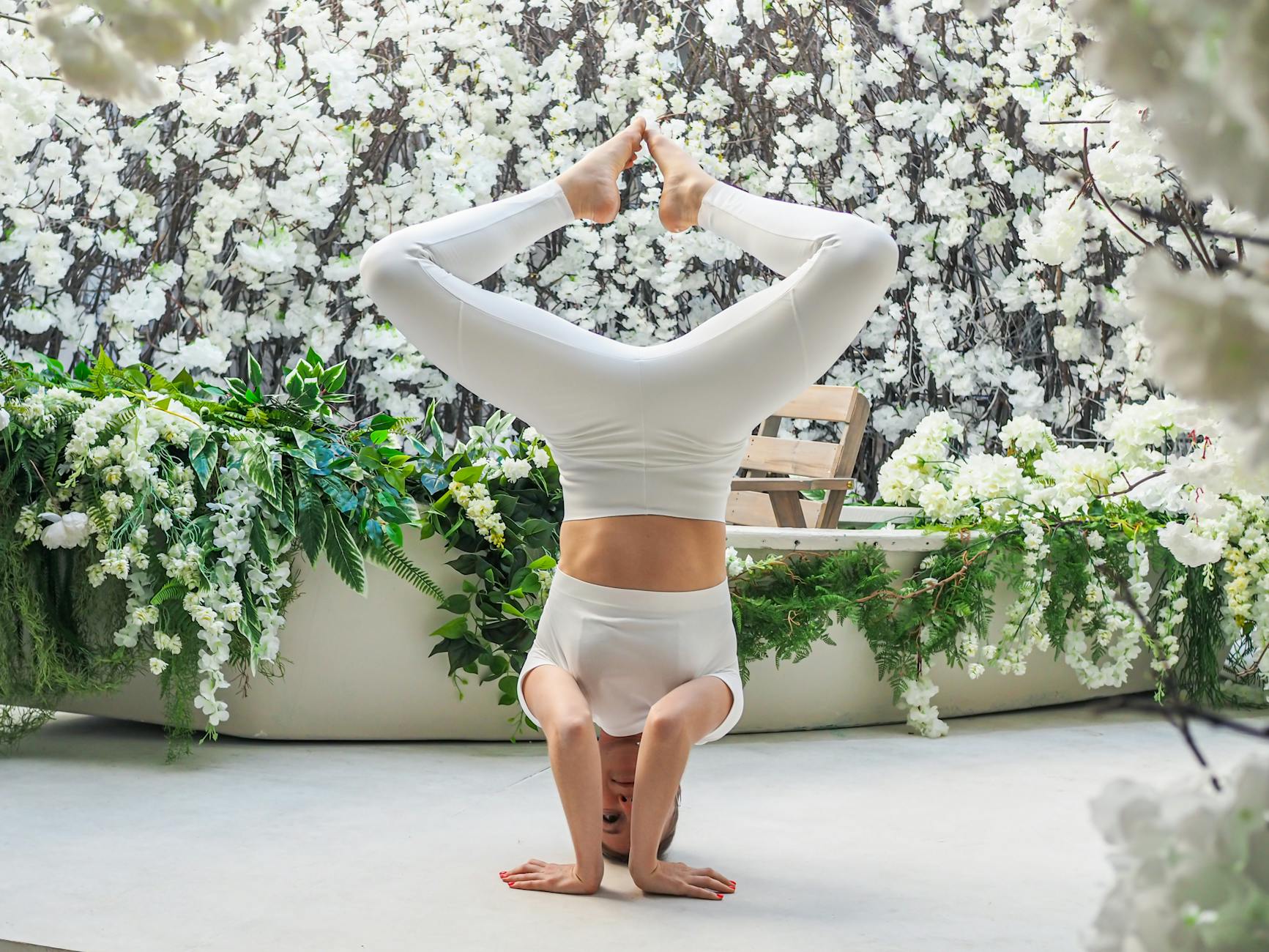 A woman performs a yoga headstand amidst a lush indoor garden of white flowers, showcasing balance and tranquility. - spring yoga renewal