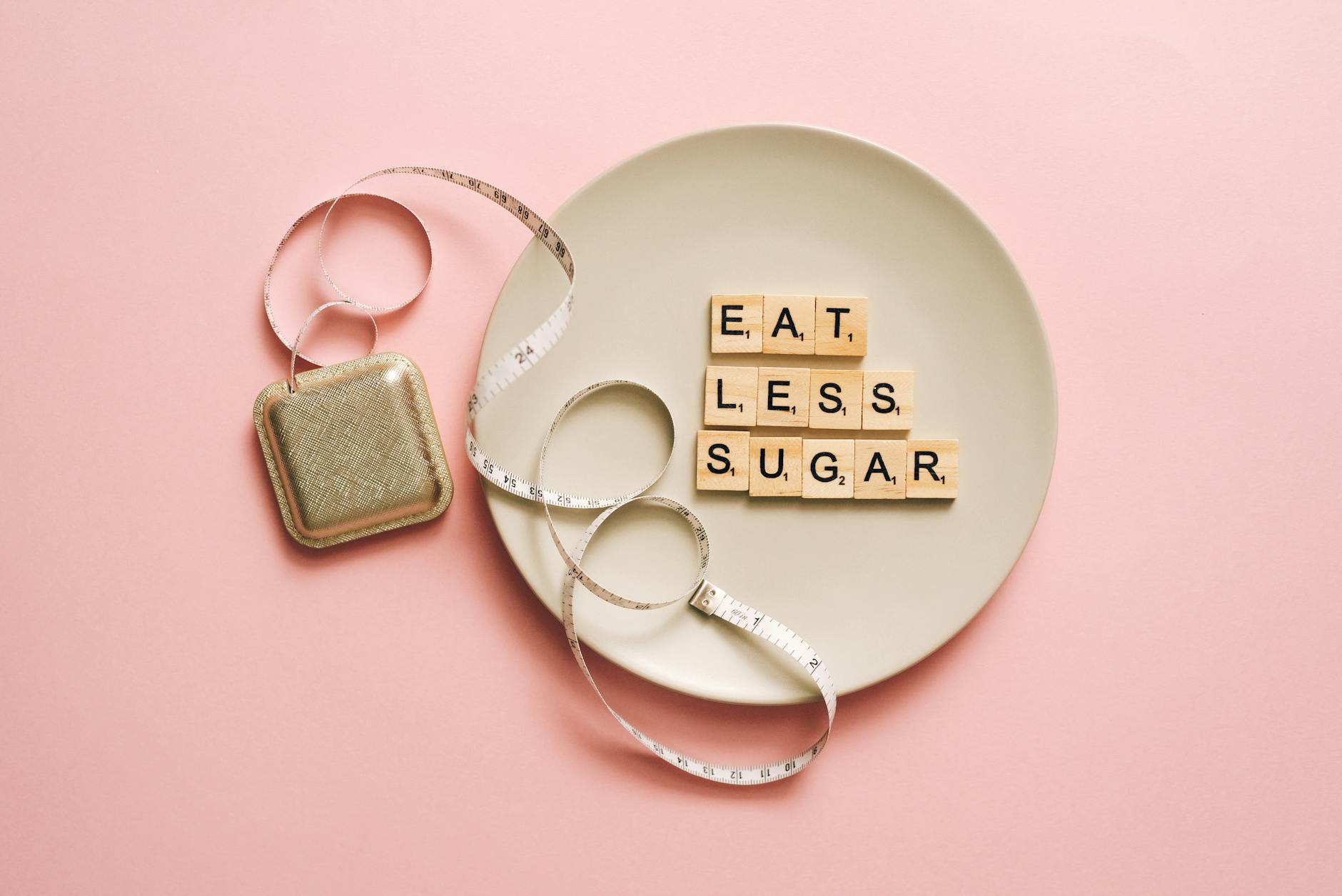 Flat lay of Scrabble tiles saying 'Eat Less Sugar' on a plate with a tape measure on a pink background. - weight loss motivation