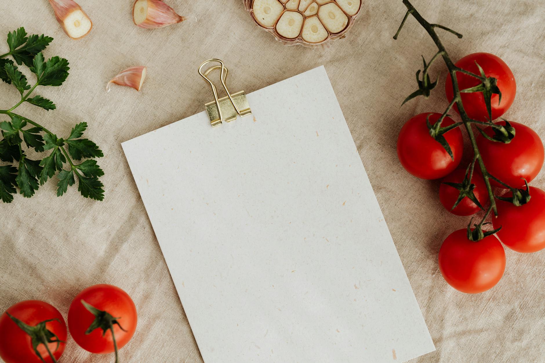 From above of blank clipboard with golden paper binder placed on linen tablecloth among tasty red tomatoes on branches together with cutted garlic and green parsley devoted for recipe or menu placement - weight watchers recipes