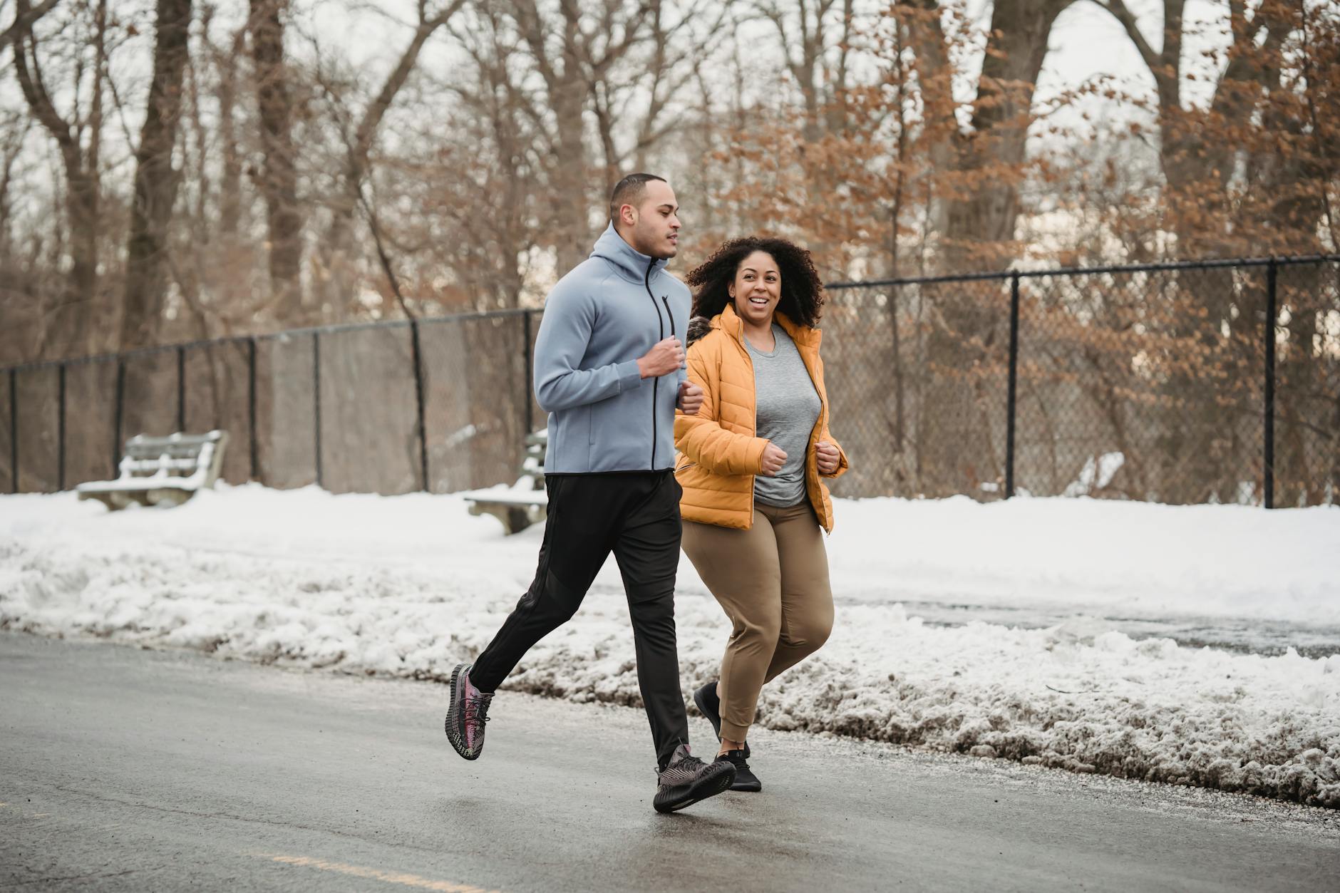 Full body content young ethnic male fitness trainer and plump black woman in warm jacket running together along snowy walkway on winter day - winter body reset
